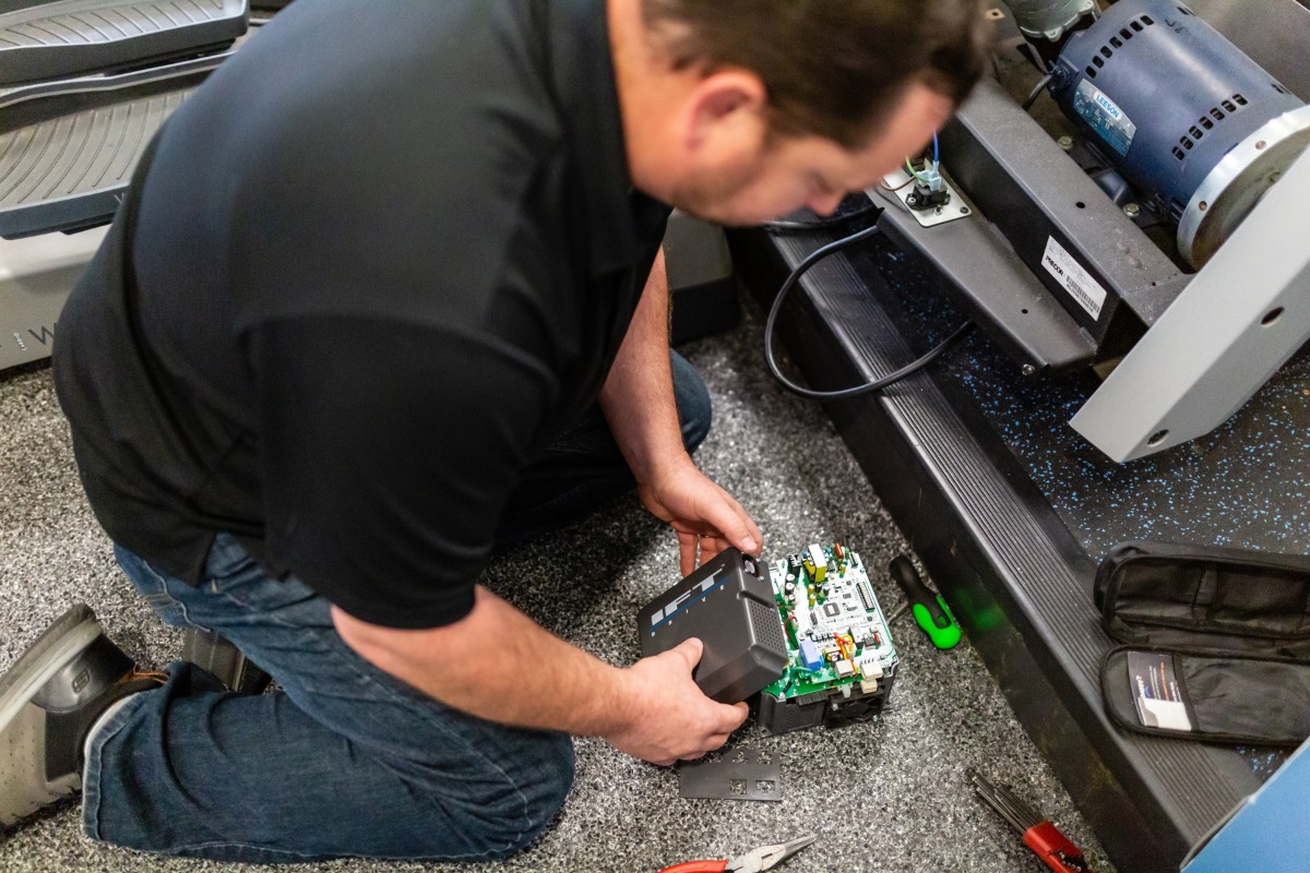 A man in a black shirt kneels on the floor, inspecting and handling an electronic device with visible circuit boards.